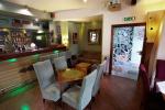 Zdjęcie przedstawia wnętrze restauracji  Szuflada Cafe&Bar, na zdjęciu widoczny drewniany stolik, obok którego stoją krzesła obite brązowym materiałem. Na stolikach leżą zielone szydełkowe serwetki. Po lewej stronie widoczny bar, a po prawej szklane drzwi, na których namalowane jest białe drzewo 