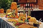 Zdjęcie przedstawia stół zastawiony jedzeniem, na którym znajdują się: tależe z owocami, ciasto mrowisko, ciasto sękacz. Z tyłu widoczny drugi stół z napojami alkoholowymi