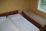 Zdjęcie przedstawia wnętrze pokoju widać dwa pojedyncze łóżka zasłane jasną pościelą stojące obok siebie. Po prawej stronie przy oknie widać pojedyncze łóżko zasłane jasnym kocem. Ściany w pokoju wykończone drewnianymi panelami.