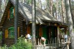 Zdjęcie przedstawia drewniany dom w lesie. Przed domem widoczny płot z patyków na którym wiszą dzbany garncarskie. Dom posiada charakterystyczne zielono - żółte okiennice. Drewniany dom znajduje się w otoczeniu lasu sosnowego