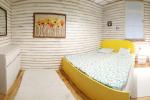 Zdjęcie przedstawia wnętrze pokoju w którym znajduje się duże łóżko małżeńskie zasłane kolorową pościelą, obicie łóżka jest w kolorze żółtym. Ściany wykończone są drewnianymi panelami, na podłodze leżą małe beżowe dywaniki. Po lewej stronie widać białą szafkę. Na ścianach widać wiszące obrazy.