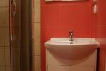 Zdjęcie przedstawia wnętrze łazienki wykończonej czerwonymi płytkami. Na zdjęciu widać białą umywalkę, szafkę oraz kabinę prysznicową. Nad umywalką znajduje się lustro. 