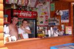 Zdjęcie przedstawia dwójkę mężczyzn stojących za drewnianym barem. Nad nimi widoczna podwieszana tablica z meni, oraz lodówka z napisem "Coca Cola". Na ladzie widoczne kubki, serwetki. 