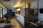 Zdjęcie przedstawia wnętrze restauracji "Żagielek", po prawej stronie widoczny zabudowany bar, na sali prostokątne stoliki, oraz krzesełka. W drugiej częsci baru widoczne czarne narożniki.   