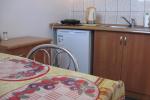 Zdjęcie przedstawia kuchnię widać: drewniane szafki kuchenne, lodówkę. Po lewej stronie widać kawałek stołu zasłanego kolorową serwetą. 
