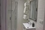  Zdjęcie przedstawia łazienkę, widać: kabinę prysznicową, zlew, lustro, grzejnik na którym wiszą ręczniki. Łazienka wykończona jest białymi płytkami.