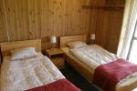 Zdjęcie przedstawia wnętrze pokoju, w którym znajdują się dwa pojedyncze łóżka zasłane jasną pościelą oraz w jednej czwartek przykryte czerwonym kocem. Obok łóżek małe szafki nocne, na których stoją lampki. Pokój wykończony jest w drewnie.