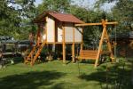 Zdjęcie przedstawia plac zabaw, widać drewniany domek stojący na belkach, oraz ławkową huśtawkę. Plac zabaw znajduje się na podwórku z przystrzyżoną trawą.