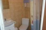 Zdjęcie przedstawia łazienkę. Widać toaletę, prysznic z zasłoną, zlew oraz białą szafkę pod zlewem. Łazienka wykończona jest beżowymi płytkami