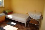 Zdjęcie przedstawia wnętrze pokoju, w którym znajduje się pojedyncze łóżko, stolik z pojedynczym krzesłem. Obok łóżka szafka nocna.