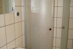 Zdjęcie przedstawia wnętrze łazienki, ściany pokrywają jasno beżowe kafelki. Na zdjęciu widoczna jest mała umywalka, nad którą jest lustro oraz kabina prysznicowa.