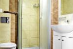 Zdjęcie przedstawia wnętrze łazienki, widać kabinę prysznicową, toaletę, zlew, lustro oraz białą szafkę. Łazienka wykończona beżowymi oraz jasnobrązowymi kafelkami